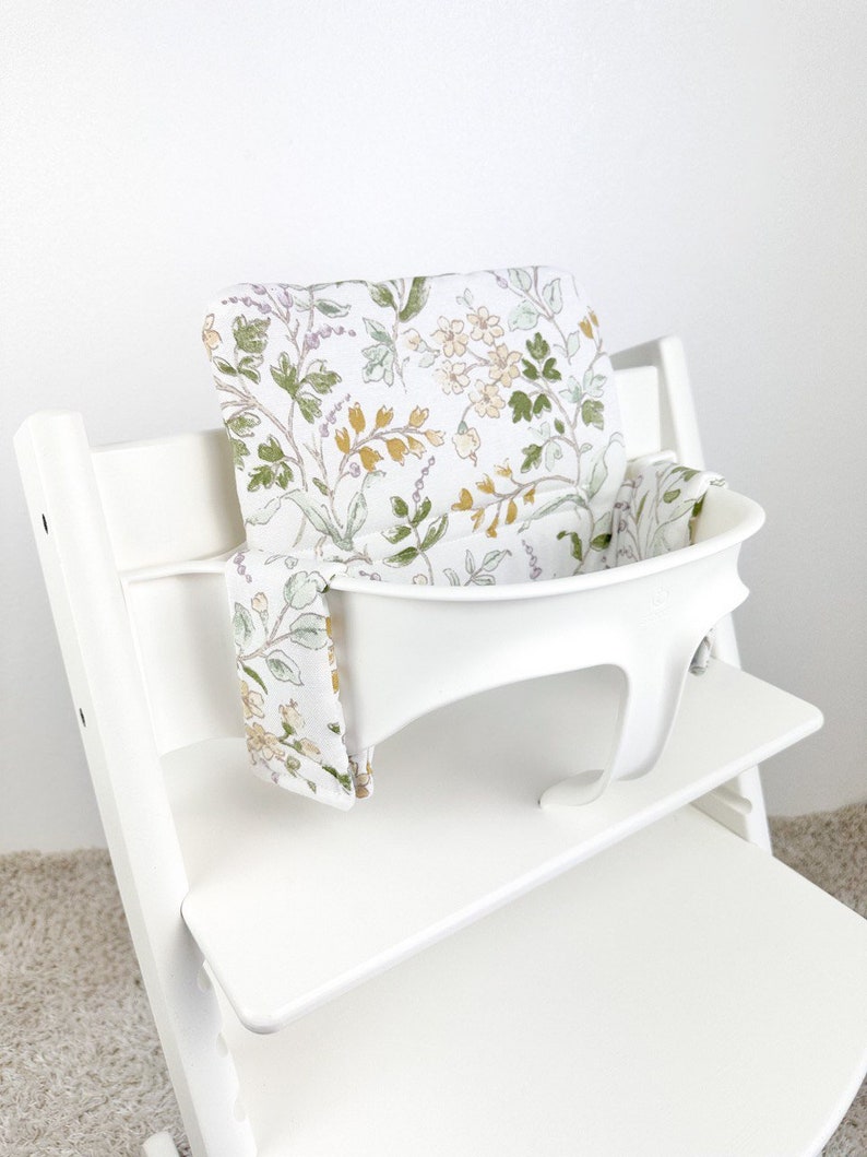 Puede incluir: Silla alta blanca con un coj&iacute;n de asiento estampado floral y una correa a juego. El coj&iacute;n tiene un fondo blanco con hojas verdes, flores amarillas y detalles morados.