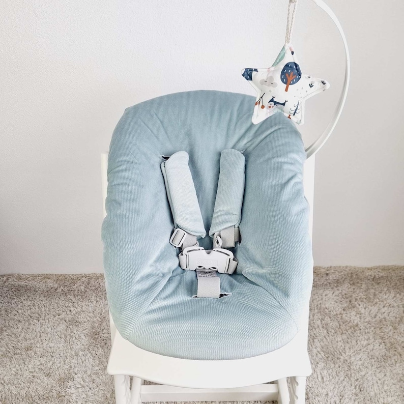 May include: A light blue baby high chair cushion with a five-point harness. A star-shaped mobile with a woodland pattern hangs above the chair. The chair is white and sits on a light-colored rug.