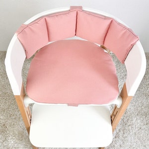May include: A pink padded high chair seat with a white frame and wooden legs. The seat has a round cushion and a padded back.