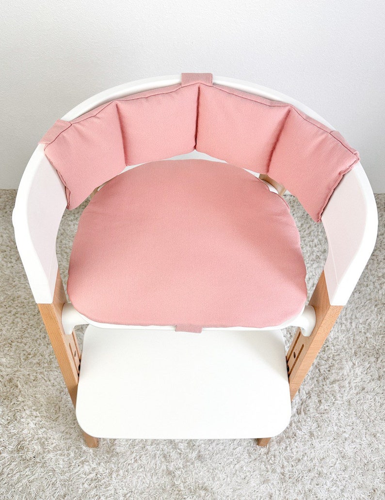 May include: A pink padded high chair seat with a white frame and wooden legs. The seat has a round cushion and a padded back.
