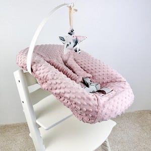 May include: A pink baby seat cushion with a textured surface, secured with gray straps, sits on a white high chair. A white arch holds a star-shaped mobile with a floral pattern.