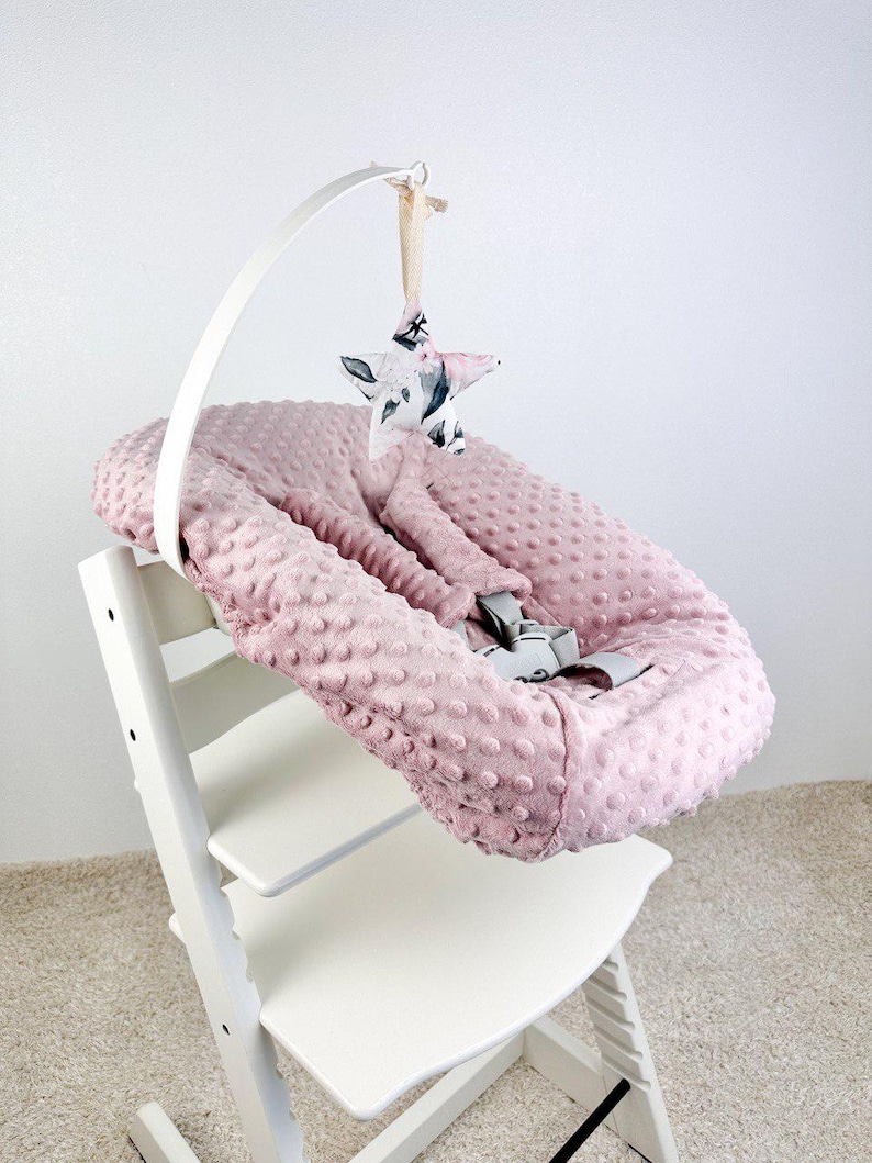 May include: A pink baby seat cushion with a textured surface, secured with gray straps, sits on a white high chair. A white arch holds a star-shaped mobile with a floral pattern.