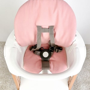 May include: A pink high chair cushion with a gray five-point harness. The cushion is made of a soft, breathable fabric and is designed to provide comfort and support for babies and toddlers.