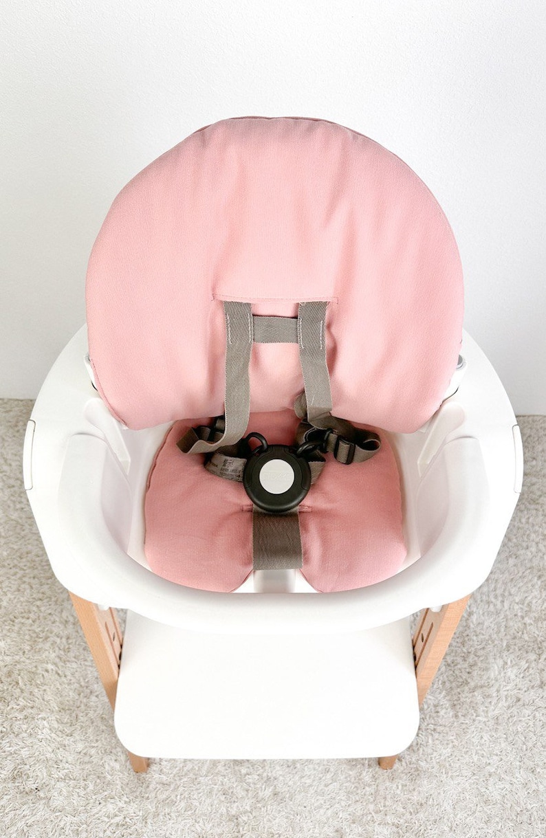 May include: A pink high chair cushion with a gray five-point harness. The cushion is made of a soft, breathable fabric and is designed to provide comfort and support for babies and toddlers.
