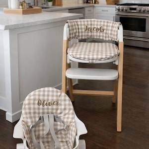 May include: A wooden high chair with a white seat and a tan and white checkered fabric cover. The cover is embroidered with the name "Jacob". A matching infant seat with the name "Oliver" is in the foreground.