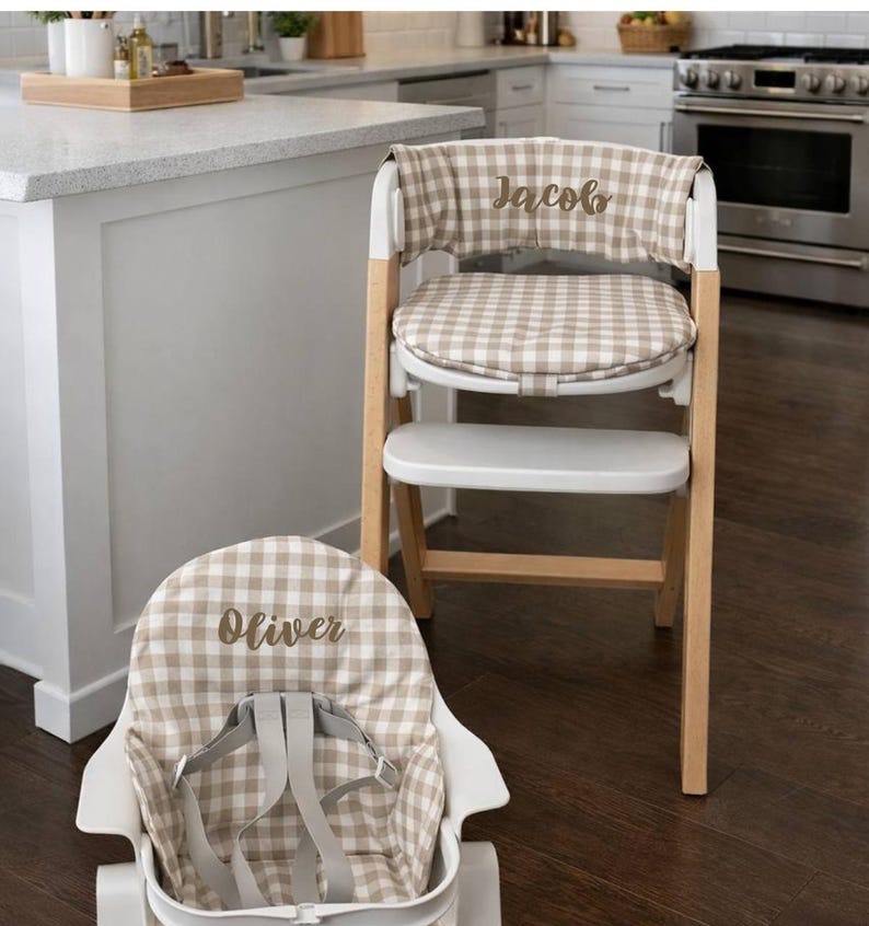 May include: A wooden high chair with a white seat and a tan and white checkered fabric cover. The cover is embroidered with the name "Jacob". A matching infant seat with the name "Oliver" is in the foreground.