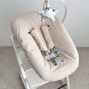 May include: A beige high chair with a star-shaped toy hanging from the top. The chair has a padded seat and backrest, with safety straps. The star toy is white with a floral pattern and green accents.