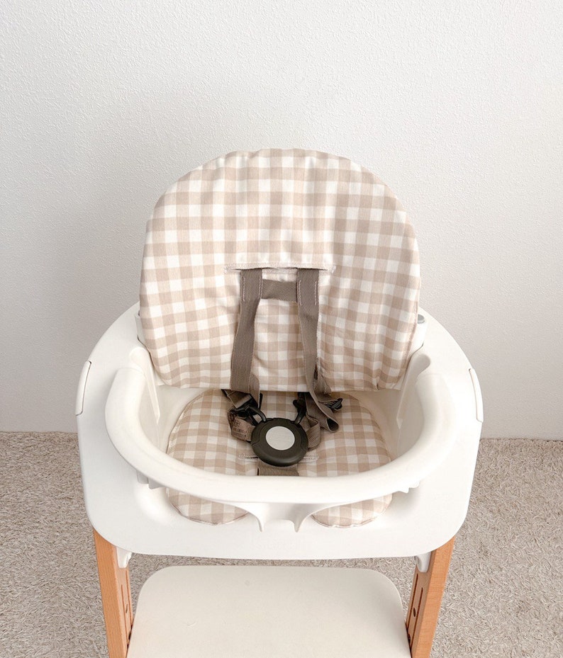 K&ouml;nnte beinhalten: Ein wei&szlig;er Hochstuhl mit einer Sitzbezug in beige-wei&szlig;em Gingham-Muster. Der Stuhl hat eine Holzbasis und einen F&uuml;nf-Punkt-Gurt.