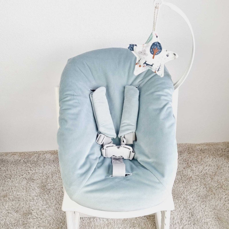 May include: A light blue baby seat cushion with a five-point harness. A star-shaped mobile with a forest pattern hangs above the seat. The seat is on a white chair.