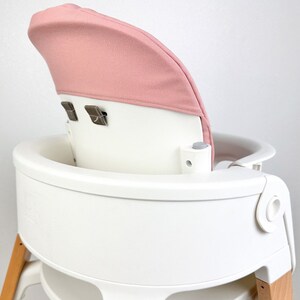 May include: A white high chair with wooden legs and a pink fabric seat back. The chair has a tray and a five-point harness.