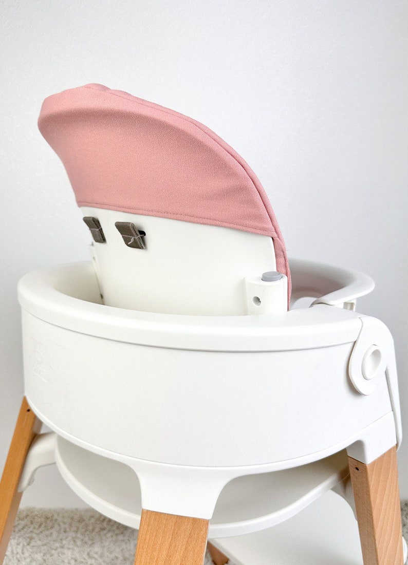 May include: A white high chair with wooden legs and a pink fabric seat back. The chair has a tray and a five-point harness.
