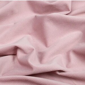 May include: Close-up of a textured, pink fabric with a woven pattern. The fabric is draped and folded, creating shadows and highlights that emphasize its texture. The color is a soft, muted pink.