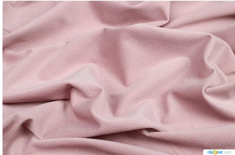 May include: Close-up of a textured, pink fabric with a woven pattern. The fabric is draped and folded, creating shadows and highlights that emphasize its texture. The color is a soft, muted pink.