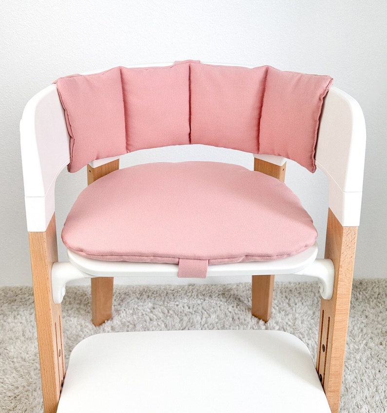 May include: A pink padded seat and back cushion for a high chair. The cushion is made of soft fabric and has a simple design.