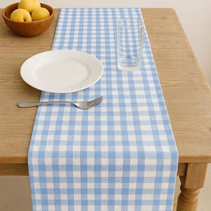 Puede incluir: Un camino de mesa a cuadros azul claro y blanco sobre una mesa de madera. Un plato blanco, un tenedor y un vaso transparente están sobre el camino de mesa. Un cuenco de madera con fruta amarilla está sobre la mesa.