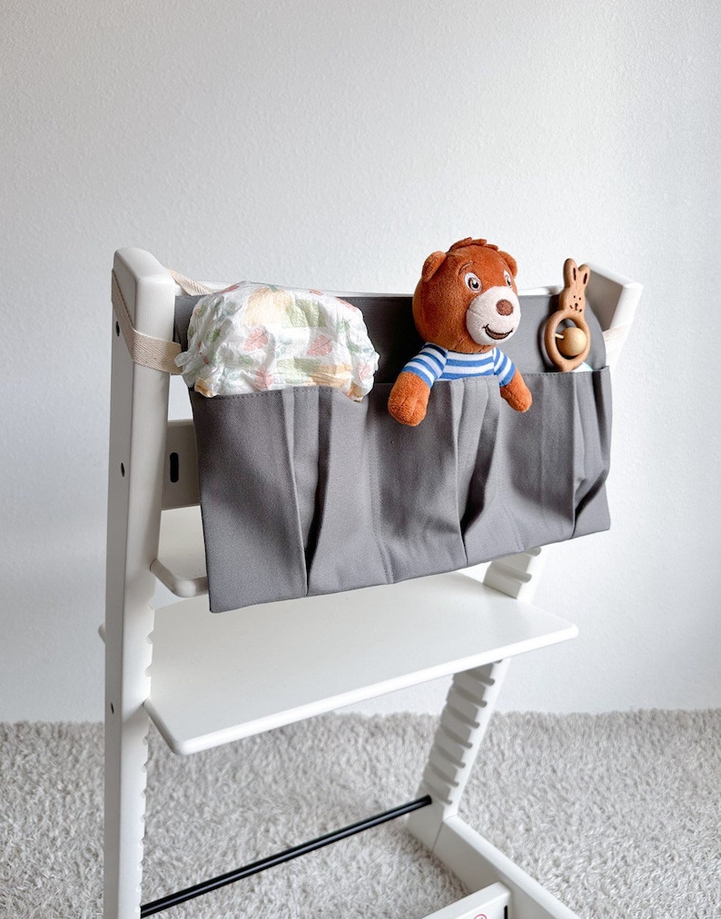 May include: Gray fabric organizer with three pockets hanging on a white high chair. The organizer has a stuffed bear, a wooden toy, and a diaper inside.