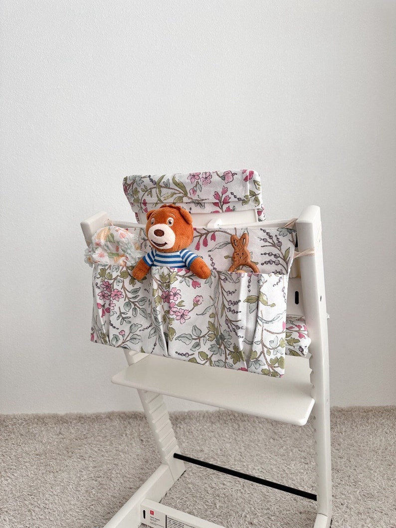 Puede incluir: Una silla alta blanca con una funda de tela con estampado floral y una bolsa de almacenamiento. La bolsa tiene tres bolsillos y est&aacute; llena de juguetes de peluche, incluyendo un oso y un conejo.