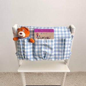 May include: A white wooden chair with a blue and white gingham fabric organizer attached to the back. A stuffed brown bear and a book with text in a foreign language are in the pockets.