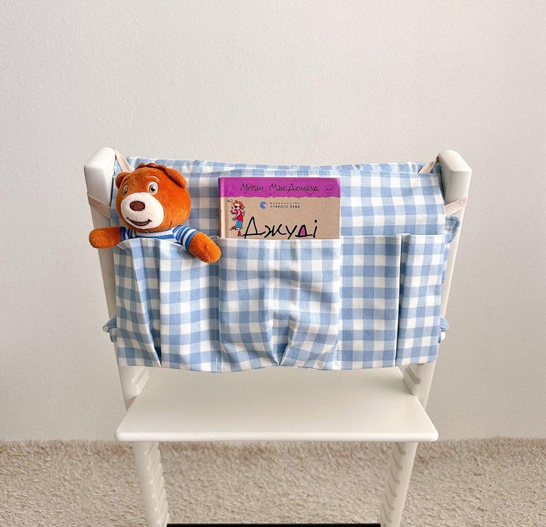 May include: A white wooden chair with a blue and white gingham fabric organizer attached to the back. A stuffed brown bear and a book with text in a foreign language are in the pockets.