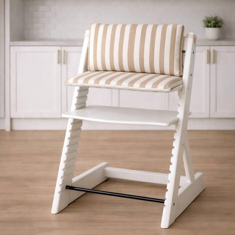 May include: A white wooden high chair with a beige and white striped cushion and backrest. The chair has a footrest and a black bar for stability. The chair is designed for a child and is placed on a wooden floor.