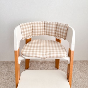 K&ouml;nnte beinhalten: Ein wei&szlig;er Hochstuhl mit einem beige-wei&szlig; karierten Kissen. Das Kissen hat eine R&uuml;ckenlehne und ein Sitzkissen.