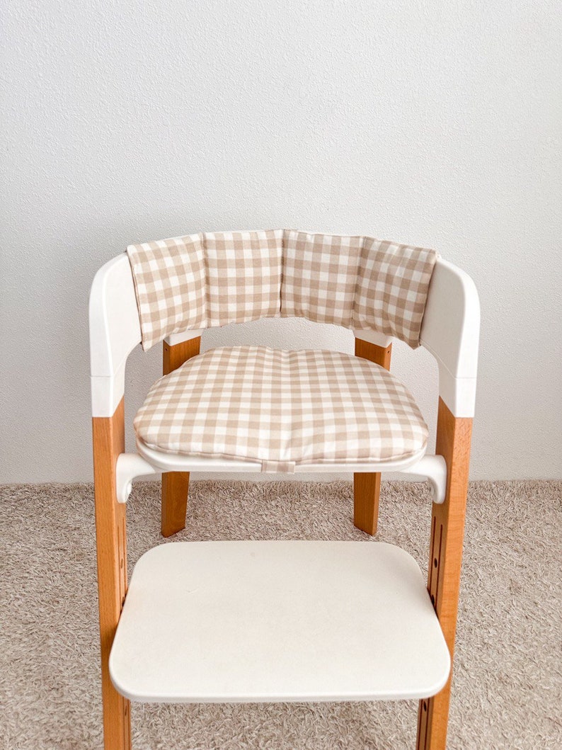 K&ouml;nnte beinhalten: Ein wei&szlig;er Hochstuhl mit einem beige-wei&szlig; karierten Kissen. Das Kissen hat eine R&uuml;ckenlehne und ein Sitzkissen.