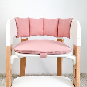 May include: A pink padded seat cushion and backrest for a high chair. The cushion is made of soft fabric and has a quilted design. The backrest is attached to the seat cushion and has a similar quilted design.