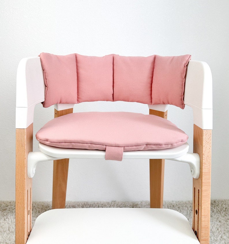 May include: A pink padded seat cushion and backrest for a high chair. The cushion is made of soft fabric and has a quilted design. The backrest is attached to the seat cushion and has a similar quilted design.