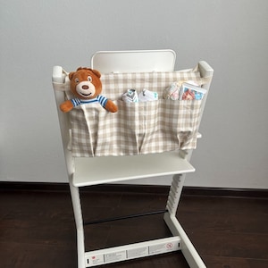 K&ouml;nnte beinhalten: Ein wei&szlig;er Hochstuhl mit einem beige-braunen karierten Stofforganizer, der an der R&uuml;ckenlehne befestigt ist. Der Organizer hat mehrere Taschen und ein Stofftier sitzt in einer der Taschen.