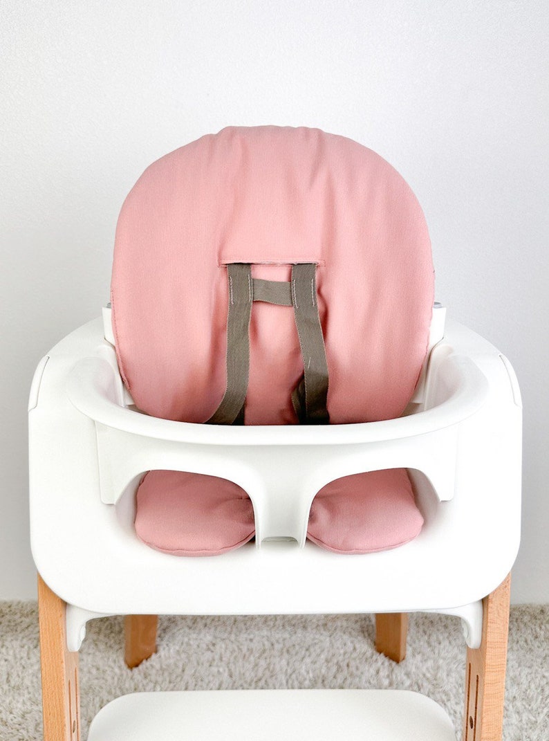 May include: A white high chair with a pink padded seat and back. The chair has a removable tray and a five-point harness.