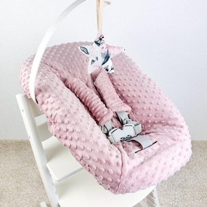 May include: A pink baby high chair cushion with a textured, dotted surface. The cushion has a safety harness and is attached to a white high chair. A star-shaped mobile with a floral pattern hangs above.