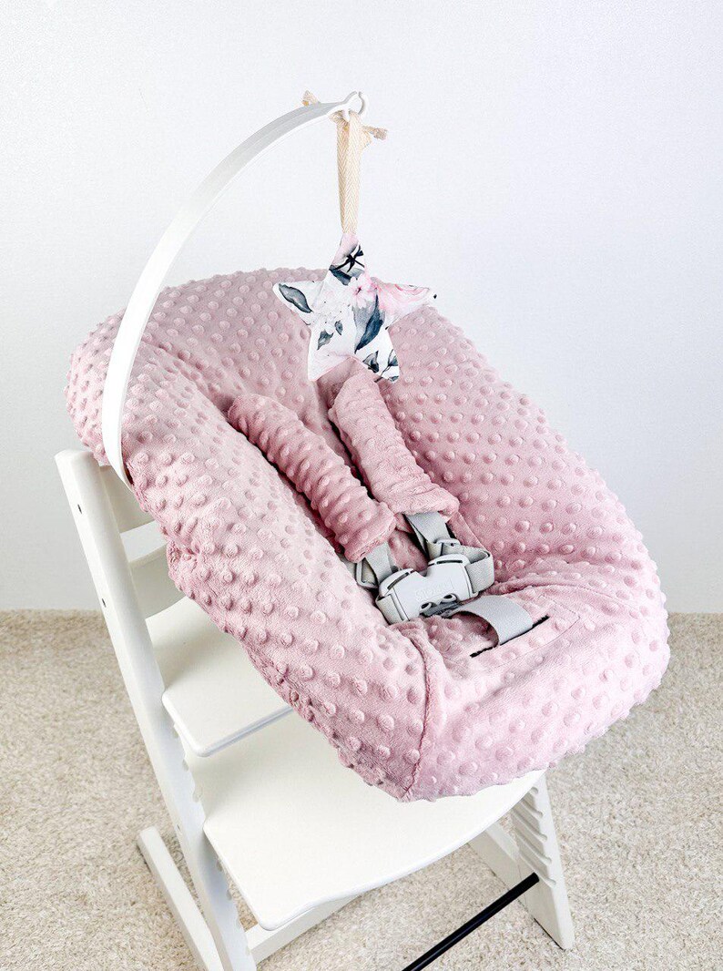 May include: A pink baby high chair cushion with a textured, dotted surface. The cushion has a safety harness and is attached to a white high chair. A star-shaped mobile with a floral pattern hangs above.