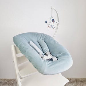 May include: A light blue baby high chair seat with a matching headrest and safety straps. A white mobile with a star-shaped fabric toy with a forest scene hangs above the seat. The high chair is white.
