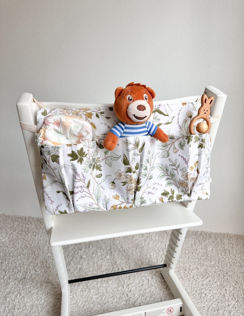 Puede incluir: Una silla alta blanca con un organizador de tela floral sujeto a la parte posterior. El organizador tiene varios bolsillos y est&aacute; lleno de un oso de peluche, un juguete de madera y un pa&ntilde;al.