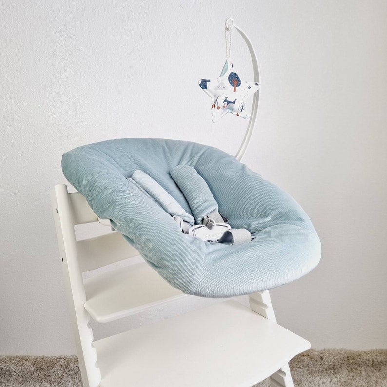 May include: A light blue baby seat with a safety harness, mounted on a white wooden high chair. A white metal arch holds a star-shaped mobile with a forest pattern. The seat is designed for infant comfort and safety.