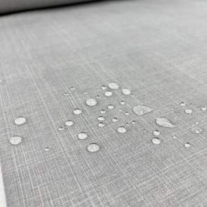 May include: Close-up of a roll of gray fabric with a textured surface. Water droplets are scattered across the fabric, indicating a waterproof or water-resistant quality. The fabric has a subtle, woven pattern.