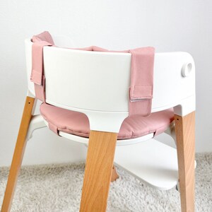 May include: A white high chair with wooden legs and a pink padded seat and straps.