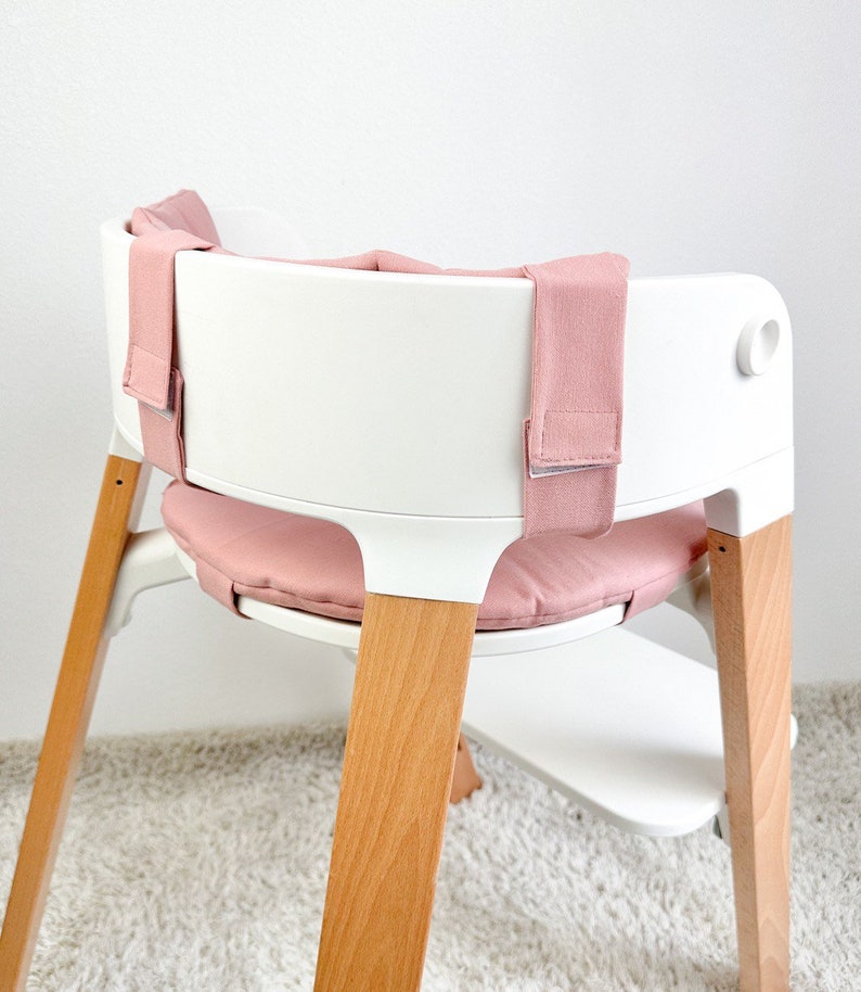 May include: A white high chair with wooden legs and a pink padded seat and straps.