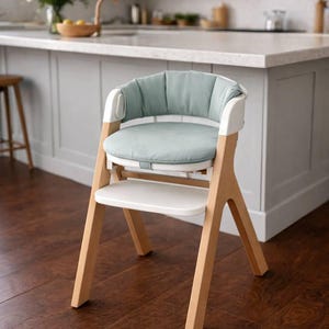 May include: A modern high chair with a light wood frame, white accents, and a soft, light blue seat cushion. The chair has a white tray and is designed for a baby or toddler. The chair is set on a dark wood floor.