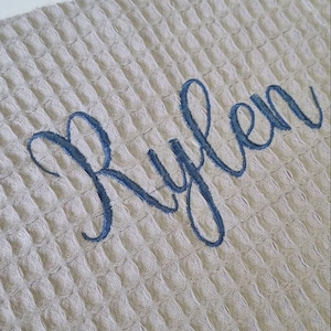 May include: A light beige waffle-weave fabric with the name "Rylen" embroidered in a flowing, dark blue script. The fabric has a textured, grid-like pattern, and the embroidery is centered on the material.