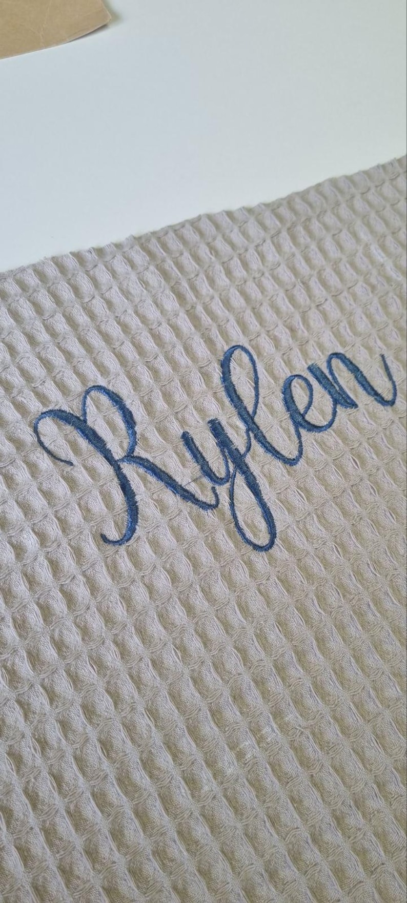 May include: A light beige waffle-weave fabric with the name "Rylen" embroidered in a flowing, dark blue script. The fabric has a textured, grid-like pattern, and the embroidery is centered on the material.