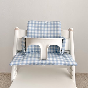 May include: A white high chair with a blue and white gingham patterned cushion and backrest. The chair has a white tray and safety straps. The chair is designed for a baby or toddler.