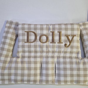 May include: A beige and white checkered pillow with the name "Dolly" embroidered in gold thread. The pillow has two side flaps and three vertical slits. The fabric has a classic gingham pattern.