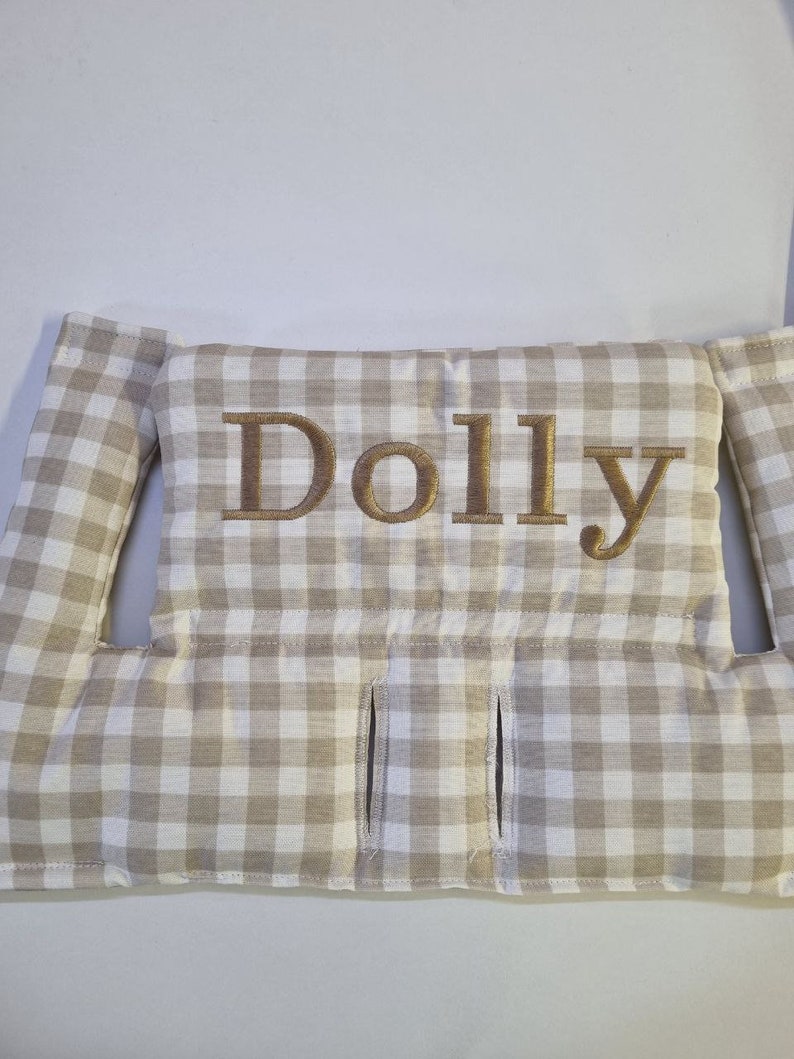 May include: A beige and white checkered pillow with the name "Dolly" embroidered in gold thread. The pillow has two side flaps and three vertical slits. The fabric has a classic gingham pattern.