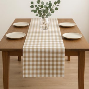Puede incluir: Una mesa de comedor de madera con un camino de mesa a cuadros beige y blanco. Cuatro platos de color crema están colocados sobre la mesa. Un jarrón de cristal transparente con ramas verdes añade un toque decorativo natural.