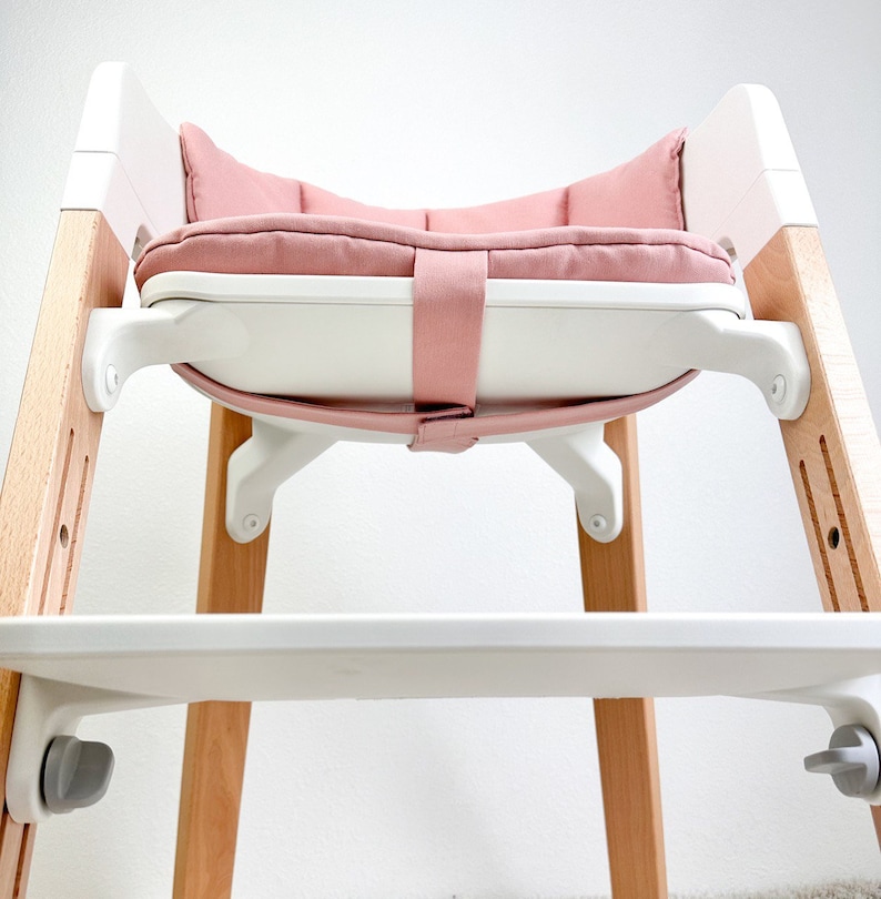 May include: A white and wooden high chair with a pink padded seat. The chair has a white tray and a safety strap.