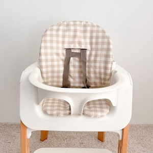 K&ouml;nnte beinhalten: Ein wei&szlig;er Hochstuhl mit einer Sitzbezug in beige-wei&szlig;em Gingham-Muster. Der Stuhl hat eine Holzbasis und ein wei&szlig;es Tablett.