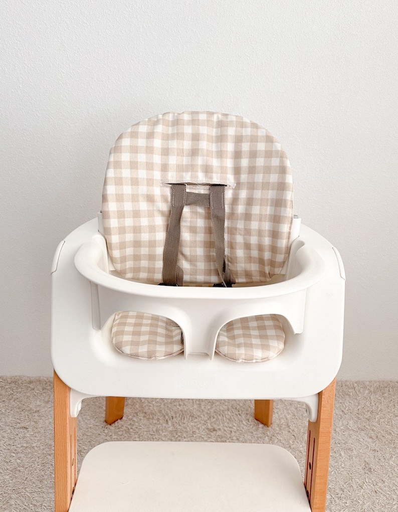K&ouml;nnte beinhalten: Ein wei&szlig;er Hochstuhl mit einer Sitzbezug in beige-wei&szlig;em Gingham-Muster. Der Stuhl hat eine Holzbasis und ein wei&szlig;es Tablett.