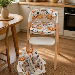 May include: A wooden high chair with a white seat and a removable pumpkin-patterned cushion. A matching infant seat is on the floor. The chair has a natural wood frame and is set in a kitchen with a round wooden table.