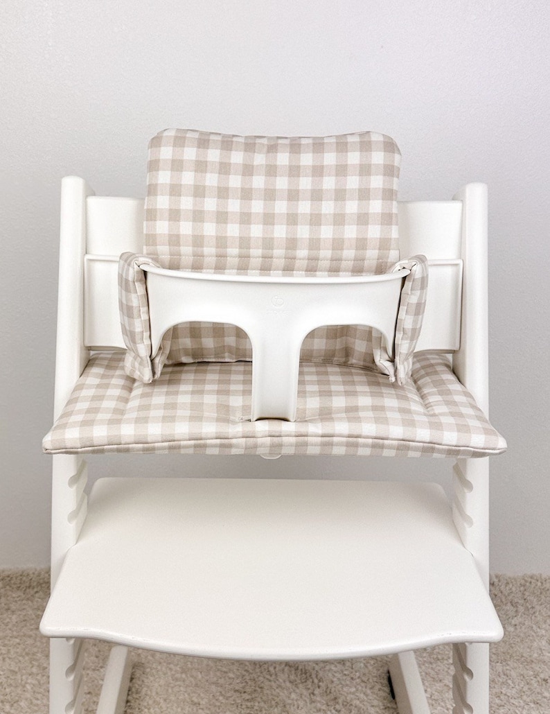 Peut inclure: Chaise haute blanche avec un coussin &agrave; carreaux beige et blanc et un plateau blanc. Le coussin a un dossier et un si&egrave;ge rembourr&eacute;s.